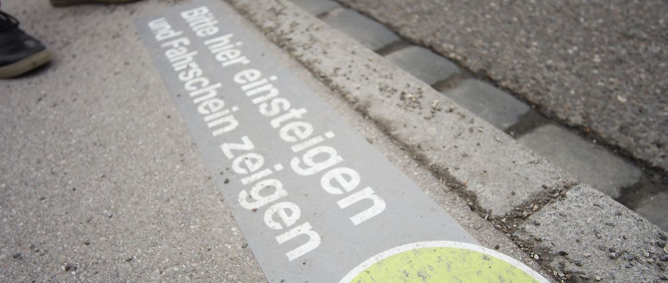 Eine Markierung am Bordstein weist mit einem Daumen-hoch-Symbol und dem Text "Bitte hier einsteigen und Fahrschein zeigen" auf eine Haltestelle hin.