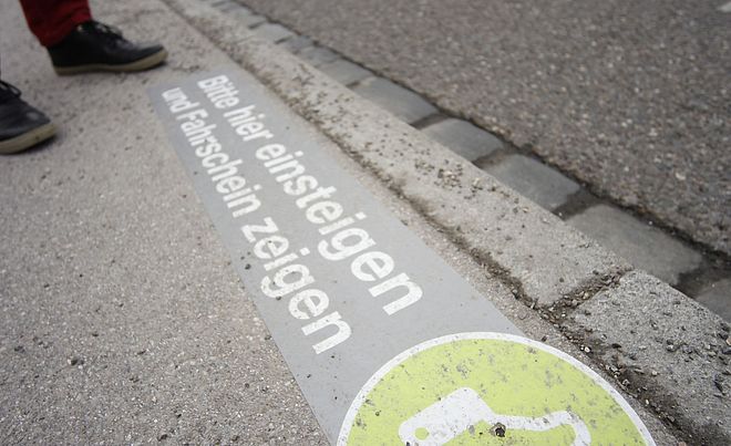 Eine Markierung am Bordstein weist mit einem Daumen-hoch-Symbol und dem Text "Bitte hier einsteigen und Fahrschein zeigen" auf eine Haltestelle hin.