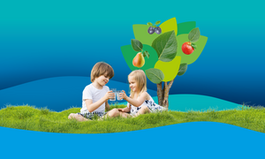 Zwei Kinder sitzen lächelnd mit einem Glas Wasser auf einer grünen Wiese. Im Hintergrund ist ein Baum mit Äpfeln