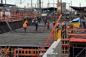 Eine Baustelle im Gleisbereich des Augsburger Hauptbahnhofs zeigt mehrere Bauarbeiter, die auf einer mit Stahlbewehrung ausgelegten Fläche arbeiten. Im Hintergrund sind Bahnanlagen und weitere Kräne zu sehen, was auf ein umfangreiches Infrastrukturprojekt hinweist, vermutlich den Bau des neuen Straßenbahntunnels. Ein Klick auf das Bild führt zu einer größeren Ansicht.