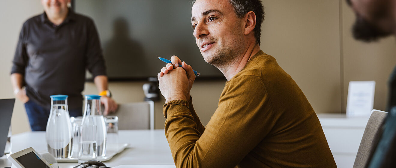 Mann sitzt mit Stift in der Hand an einem Konferenztisch. Im Hintergrund steht ein Mann vor einem Bildschirm