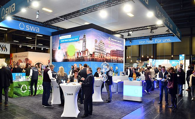 Eine belebte Veranstaltung zeigt zahlreiche Menschen, die an Stehtischen mit dem SWA-Logo sich unterhalten, während im Hintergrund ein großer beleuchteter Stand mit einer Augsburger Stadtansicht das Bild prägt.