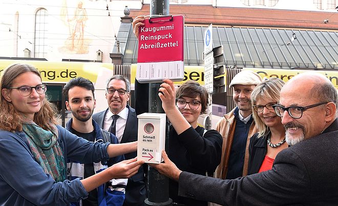 Mehrere Personen stehen an einer Stange, an welcher Abreißzettel und Kaugummi-Kästen zur Entsorgung von Kaugummis befestigt sind. Im Hintergrund befindet sich eine Straßenbahn am Moritzplatz.