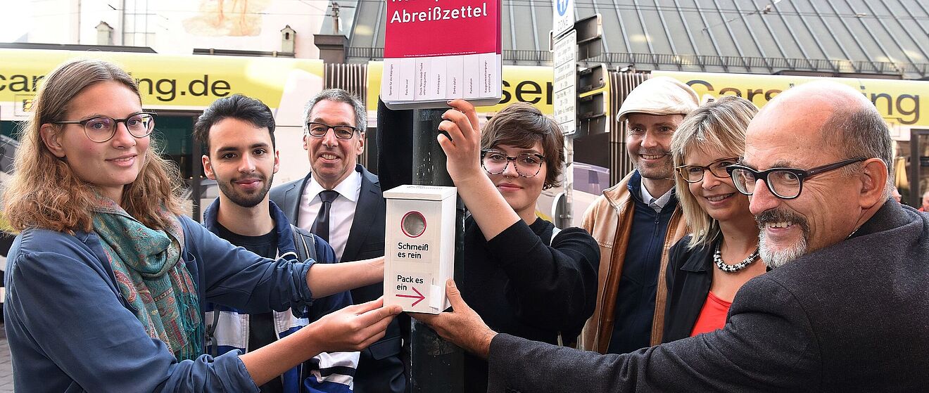 Mehrere Personen stehen an einer Stange, an welcher Abreißzettel und Kaugummi-Kästen zur Entsorgung von Kaugummis befestigt sind. Im Hintergrund befindet sich eine Straßenbahn am Moritzplatz.