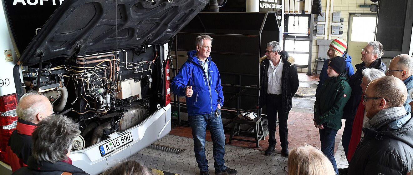 Gäste bei einem Programmpunkt in der Buswerkstatt. Ein Mitarbeiter des Busbetriebshof informiert die Gäste über Komponenten an einem Bus.