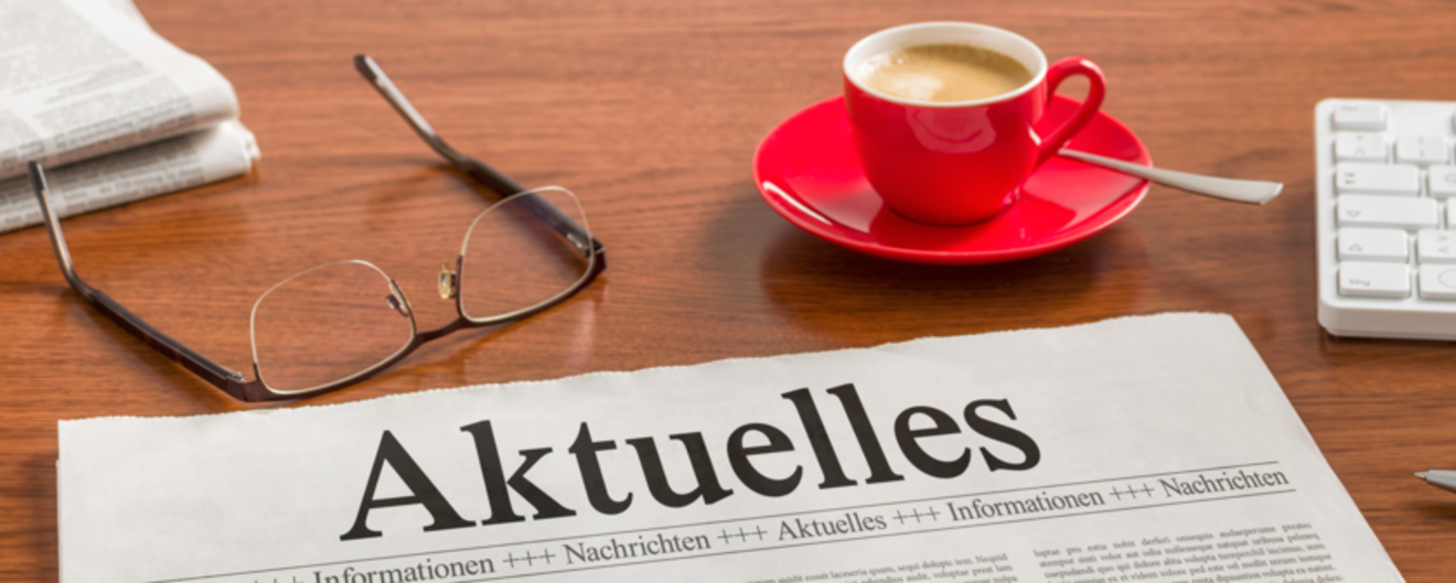 Tisch auf dem Zeitungen, eine Tastatur, eine Brille, ein Stift und eine Tasse Kaffee liegen