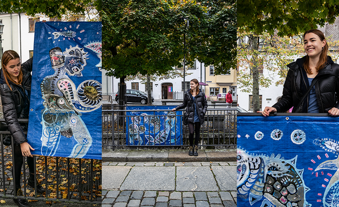Im Vordergrund trägt eine junge Frau eine schwarze Daunenjacke und Jeans, während sie auf einem gepflasterten Gehweg neben einem kunstvollen blauen Wandbehang posiert.