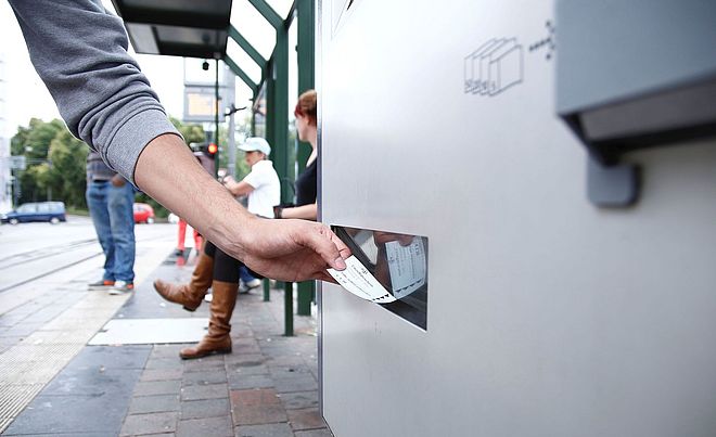 Eine Person, die an einer Haltestelle ein Ticket aus einem Fahrkartenautomaten nimmt.