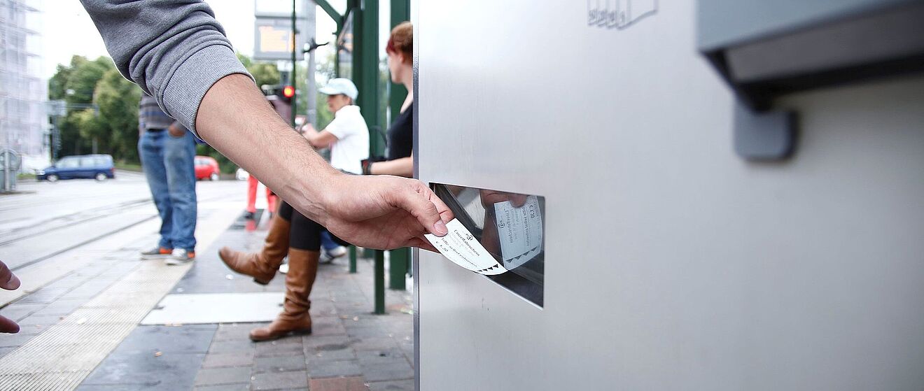Eine Person, die an einer Haltestelle ein Ticket aus einem Fahrkartenautomaten nimmt.