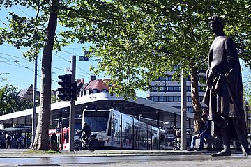 Am Königsplatz fährt eine Tram, ein Fahrradfahrer und rechts steht eine Statue. Ein Klick führt zur Vergrößerung des Bildes.