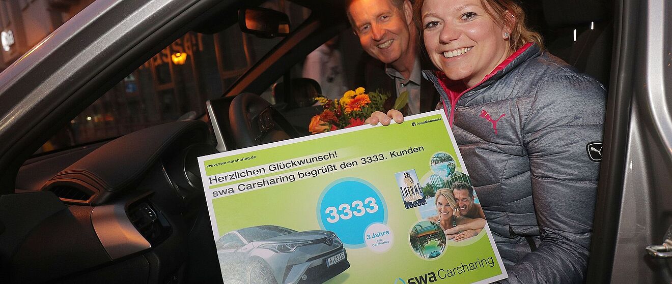 Eine Frau und ein Mann sitzen in einem Fahrzeug der swa-Carsharing-Flotte. Die Frau im Vordergrund hält einen großen Gutschein für den 3.333sten Kunden im swa-Carsharing. Im Hintergrund befindet sich der Mann mit einem Blumenstrauß.