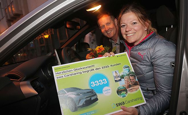 Eine Frau und ein Mann sitzen in einem Fahrzeug der swa-Carsharing-Flotte. Die Frau im Vordergrund hält einen großen Gutschein für den 3.333sten Kunden im swa-Carsharing. Im Hintergrund befindet sich der Mann mit einem Blumenstrauß.