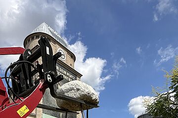 Ein massiver Stein wird von einem Gabelstapler auf Eisenstäbe gehoben. Im Hintergrund ist das Ofenhaus und der Gaskessel auf dem historischen Gaswerk in Augsburg zu erkennen.