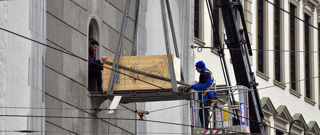 Mann auf einem Personenaufzug und ein Mann im Fenster transportieren eine Box für den Turamichele mit swa Branding.