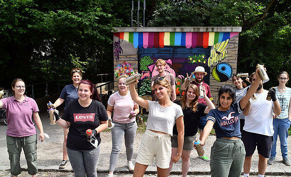 Gruppe hält Graffitidosen in den Händen vor einem bereits angesprühten Wand eines Technikgebäudes der Stadtwerke Augsburg.