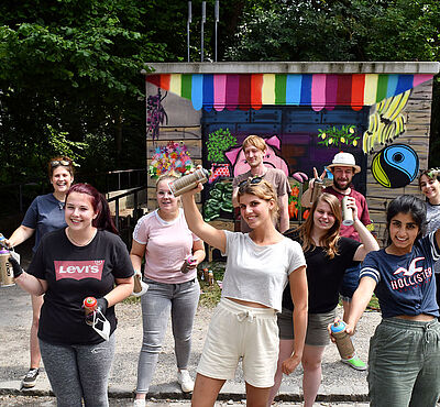 Gruppe hält Graffitidosen in den Händen vor einem bereits angesprühten Wand eines Technikgebäudes der Stadtwerke Augsburg.