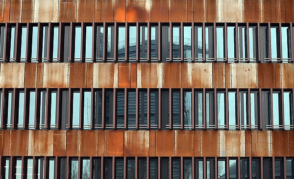 Fensterfassade eines Gebäudes in rostiger Optik auf dem Gaswerksgelände. In der Reflektion befindet sich der Gaskessel.