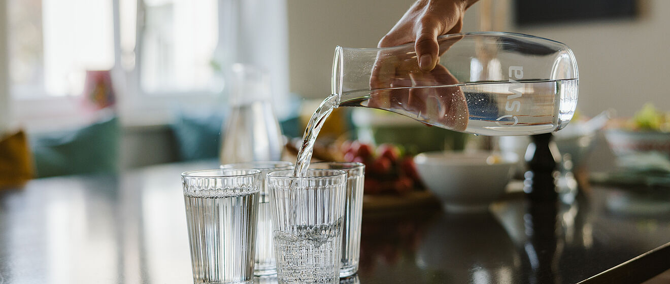 Person befüllt Gläser auf einem Tisch mit Wasser aus einer Karaffe