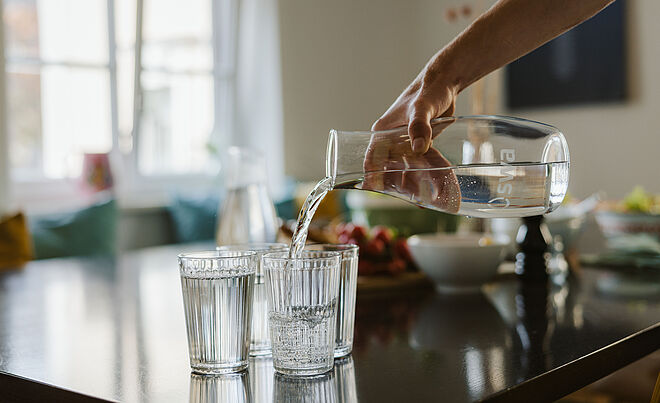 Person befüllt Gläser auf einem Tisch mit Wasser aus einer Karaffe