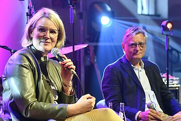 Frau mit Mikrofon sitzt vor Instrumenten. Im Hintergrund ist ein Mann mit Sakko. - Ein Klick auf das Bild führt zur vergrößerten Fotoansicht.