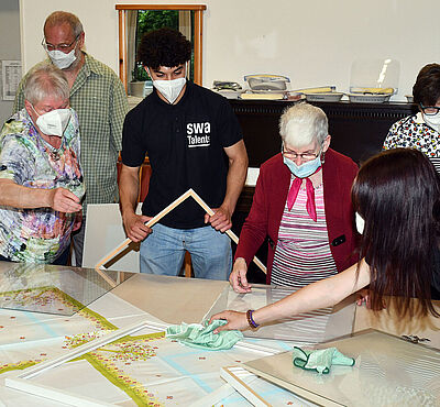 Gruppe Arbeitet zusammen, bestehend aus zwei "swa-Talents" und 5 älteren Damen und Herren. Sie tragen alle einen Mundschutz und basteln an Bilderrahmen.