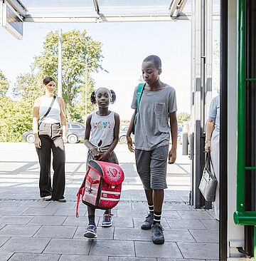 Mutter läuft mit Tochter, die eine Schultasche in der Hand hält auf einer Haltestelle