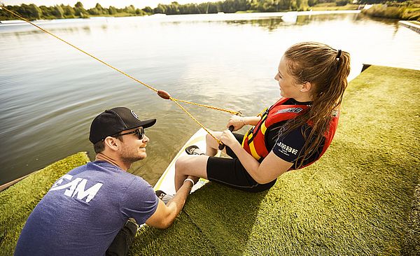 Ein Mann und eine Frau sitzen an einem See auf einem Grünstreifen. Die Frau sitzt rechts, trägt eine Schwimmweste und hält einen Griff in denen Händen, woran ein Seil befestigt ist. Der Mann sitzt  links daneben und hält ihre Beine fest.