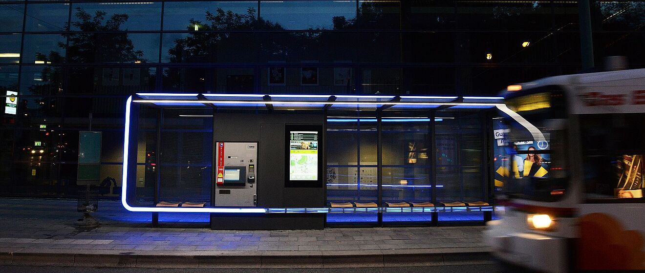 Vorne auf der rechten Seite ist die Vorderseite einer Straßenbahn. Im Hintergrund befindet sich eine Haltestelle dieser Straßenbahn.