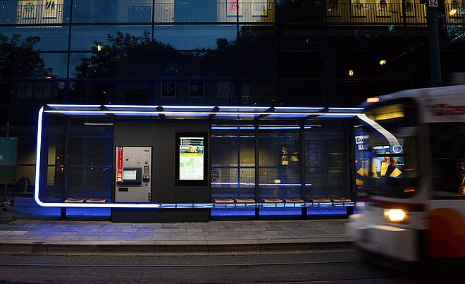 Vorne auf der rechten Seite ist die Vorderseite einer Straßenbahn. Im Hintergrund befindet sich eine Haltestelle dieser Straßenbahn.