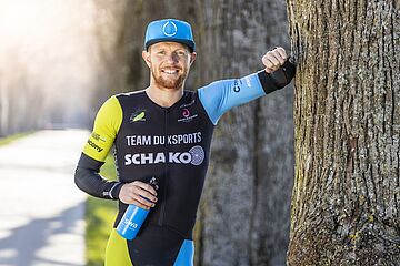 Triathlet Lukas Stahl in Sportklamotten, einer swa Kappe auf dem Kopf und einer swa Trinkflasche in der Hand stützt sich an einen Baum