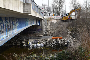 Fluss und Wertachbrücke. Im Hintergrund fährt ein Bagger einen Hang hoch. Ein Klick sorgt für eine Großansicht des Bildes.