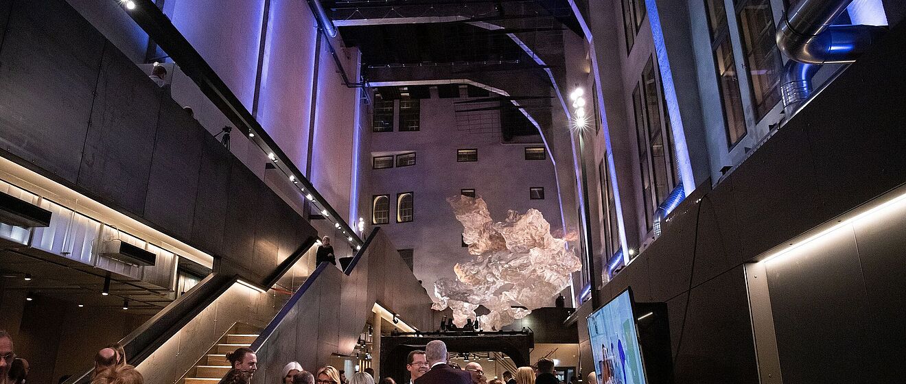 Mehrere vornehm gekleidete Personen befinden sich im historisch-industriellen Ofenhaus Augsburg. Im Hintergrund befindet sich eine Luftskulptur in Form einer Rauchwolke.