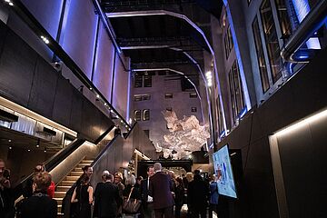 Mehrere vornehm gekleidete Personen befinden sich im historisch-industriellen Ofenhaus Augsburg. Im Hintergrund befindet sich eine Luftskulptur in Form einer Rauchwolke. - Ein Klick auf das Bild führt zur vergrößerten Fotoansicht.