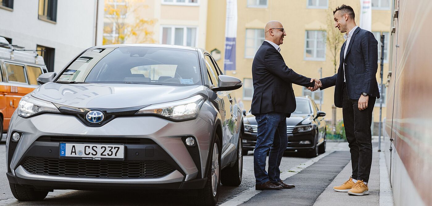 Zwei Männer im Sakko geben sich auf dem Gehweg die Hand und stehen neben einem Toyota
