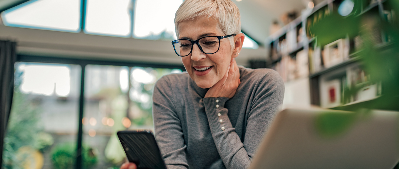 Frau mit Smartphone in der Hand steht vor einem Laptop. 