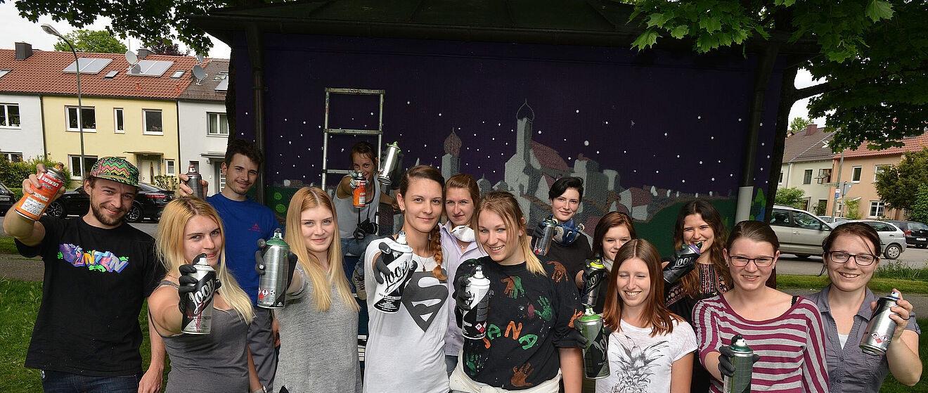 Gruppe von Menschen mit Sprühdosen in der Hand. Hinter ihnen eine Leinwand einem Kunstwerk der Stadt.