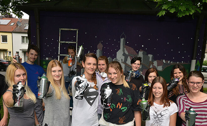 Gruppe von Menschen mit Sprühdosen in der Hand. Hinter ihnen eine Leinwand einem Kunstwerk der Stadt.