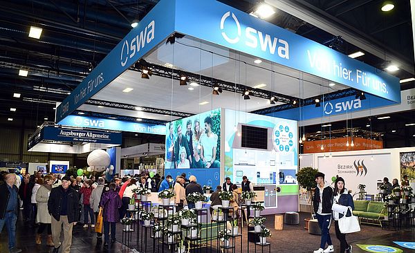  Ein großer, beleuchteter Messestand der swa zeigt zahlreiche Besucher, die sich im Bereich von Pflanzenarrangements und einem großen digitalen Bildschirm aufhalten.
