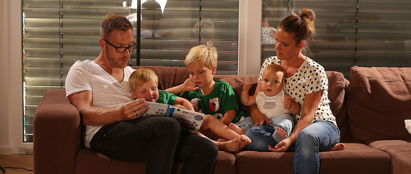 Augsburger Familie mit drei Kindern sitzt einem Sofa und liest ein Kinderbuch.