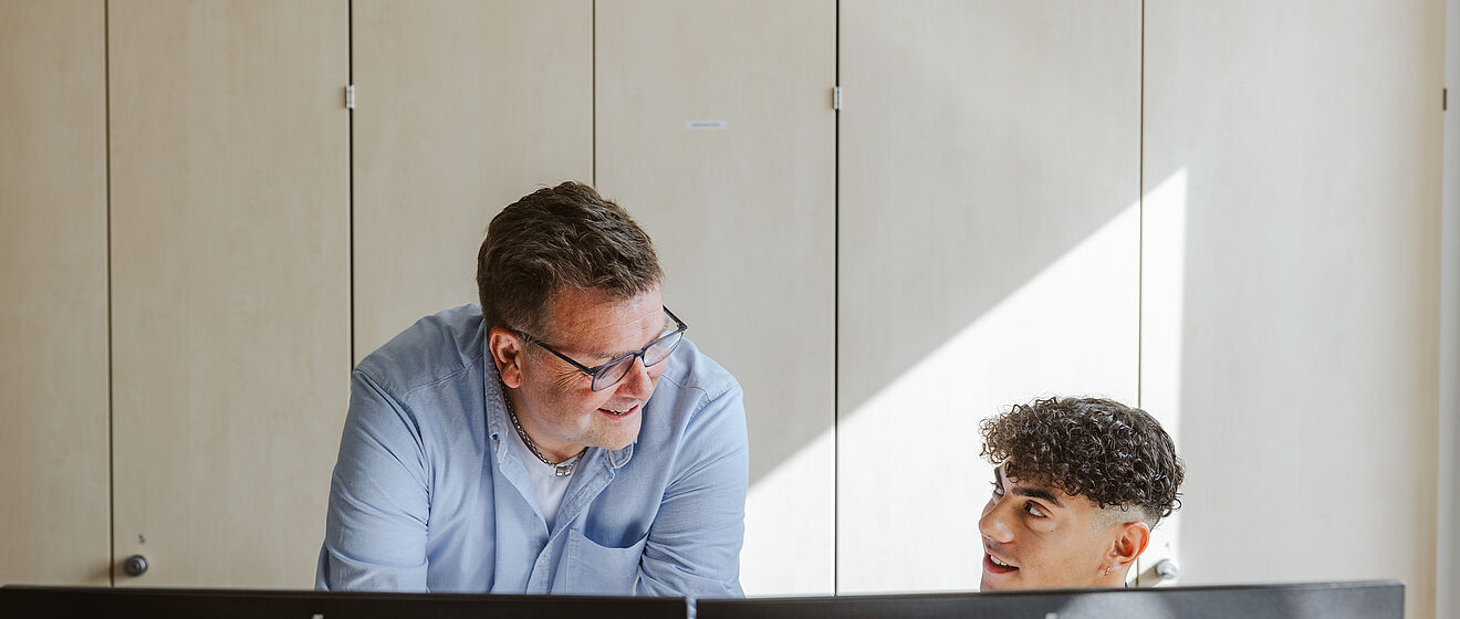 Ein Mann sitzt am Schreibtisch vor einem Monitor und unterhält sich mit einer stehenden Person neben ihm