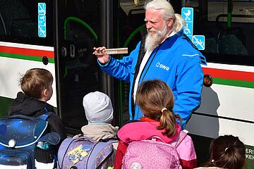 Mann mit Bart und swa Jacke erklärt Kindern etwas beim Bussicherheitstraining. Im Hintergrund ist ein Bus