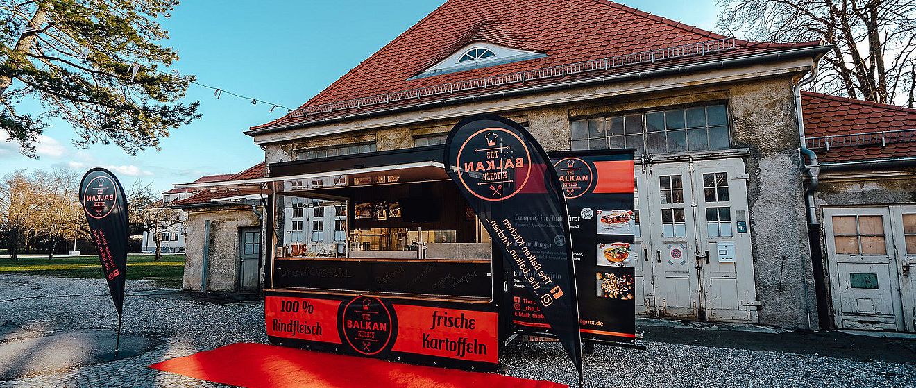 Frontansicht auf Balkan Kitchen mit Bannern und roten Teppich auf dem Boden. Weiterverlinkung auf The Balkan Kitchen Newsartikel