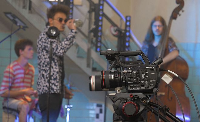 Filmkamera. Im Hintergrund sind Leute mit Instrumenten. Weiterverlinkung auf Stay Home Festival Newsartikel