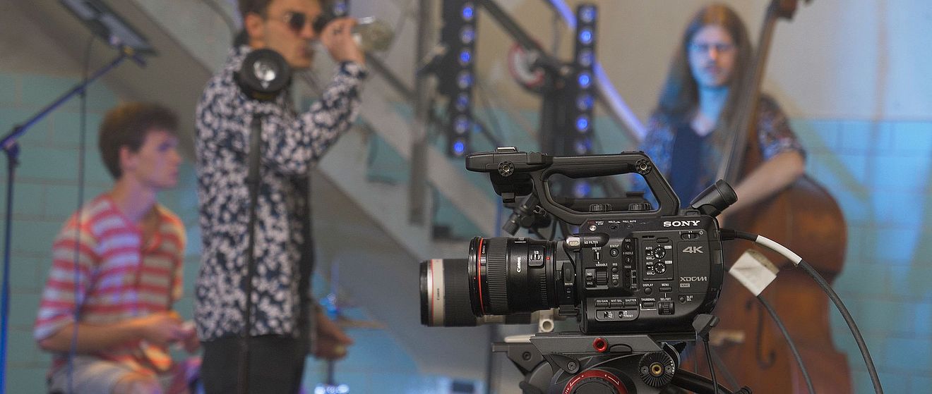 Filmkamera. Im Hintergrund sind Leute mit Instrumenten. Weiterverlinkung auf Stay Home Festival Newsartikel