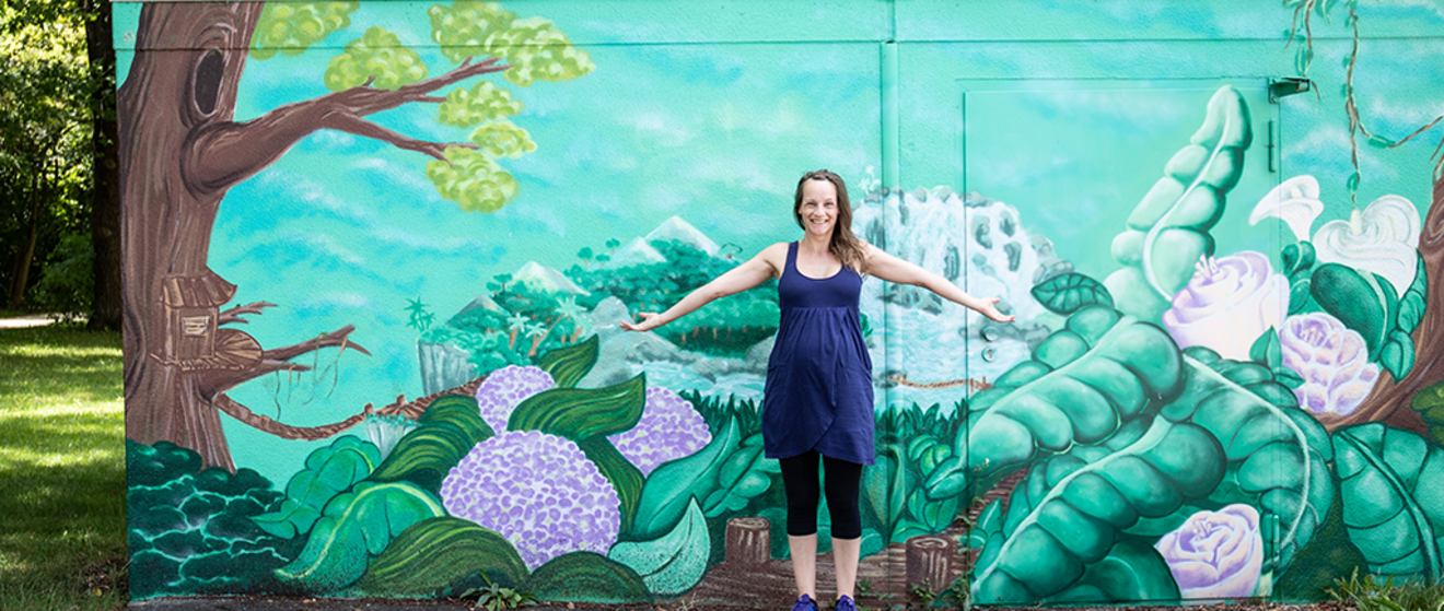 Eine Person steht mit ausgebreiteten Armen vor einem großen, farbenfrohen Wandgemälde mit floralen und landschaftlichen Motiven. Die Frau im Bild ist Sophie Te, eine Augsburger Graffiti-Künstlerin und Rapperin, die Teil des Projekts #AugsburgArtists der Stadtwerke Augsburg (swa) war.