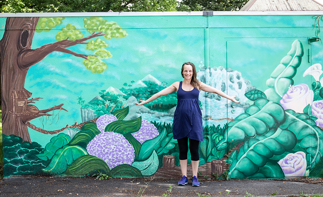 Eine Person steht mit ausgebreiteten Armen vor einem großen, farbenfrohen Wandgemälde mit floralen und landschaftlichen Motiven. Die Frau im Bild ist Sophie Te, eine Augsburger Graffiti-Künstlerin und Rapperin, die Teil des Projekts #AugsburgArtists der Stadtwerke Augsburg (swa) war.