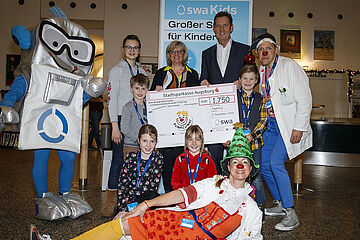 Eine Gruppe von Kindern und Erwachsenen posiert für ein Foto bei einer Spendenübergabe der Stadtsparkasse Augsburg. Der große, silberne Roboter namens Oswald (swa Maskottchen) steht links neben den Kindern und zwei Personen in Weihnachtskostümen sind ebenfalls anwesend. Ein Klick auf das Bild führt zu einer größeren Ansicht.