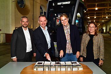 Vier Personen stehen vor einem Tisch mit einem Kuchen in Straßenbahnoptik. Einer der Männer hält ein Messer und schneidet den Kuchen an. Im Hintergrund ist eine Straßenbahn. Ein Klick sorgt für eine Großansicht des Bildes.