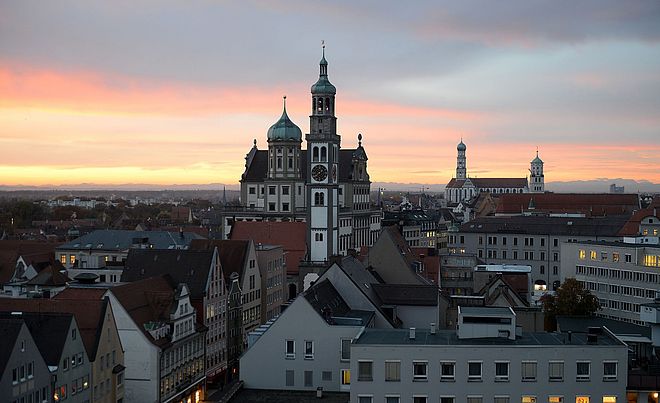 Eine Luftaufnahme zeigt die historische Altstadt von Augsburg bei Sonnenuntergang oder Sonnenaufgang, mit dem markanten Perlachturm und dem Rathaus im Zentrum der Skyline.