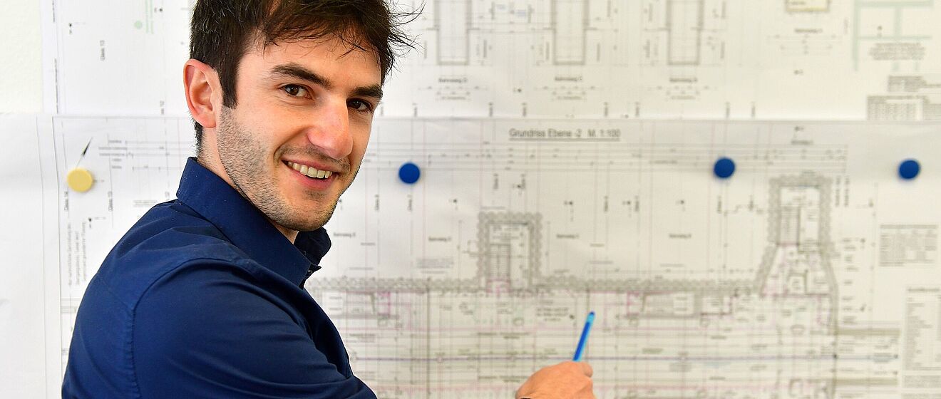 Mann zeigt mit einem Stift auf einen Papierplan an der Wand.
